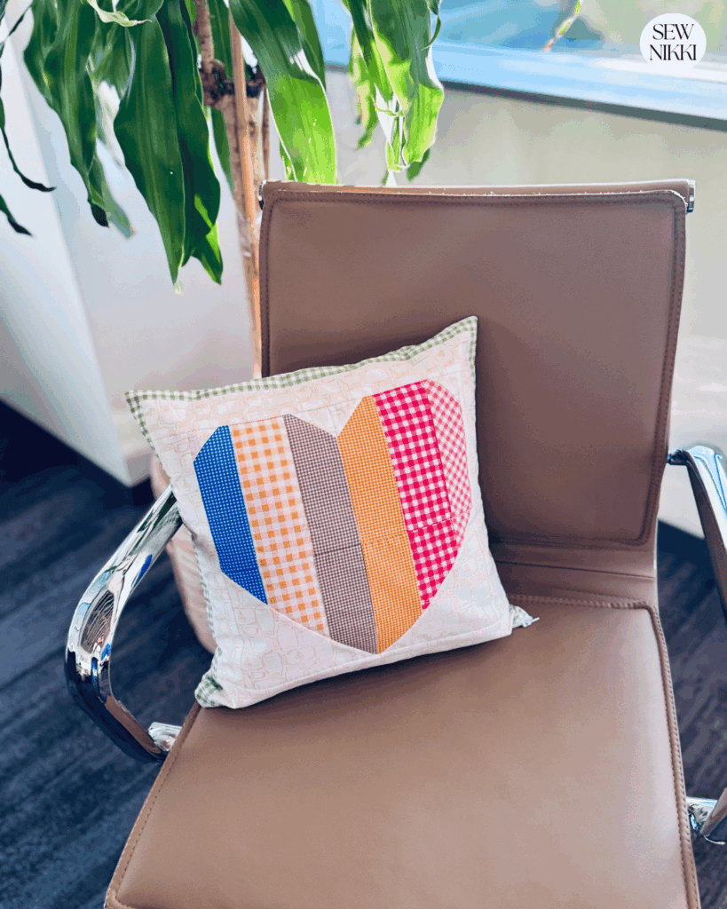 Finished Patchwork Striped Heart Block Pillow by Sew Nikki sitting on a chair next to plant.